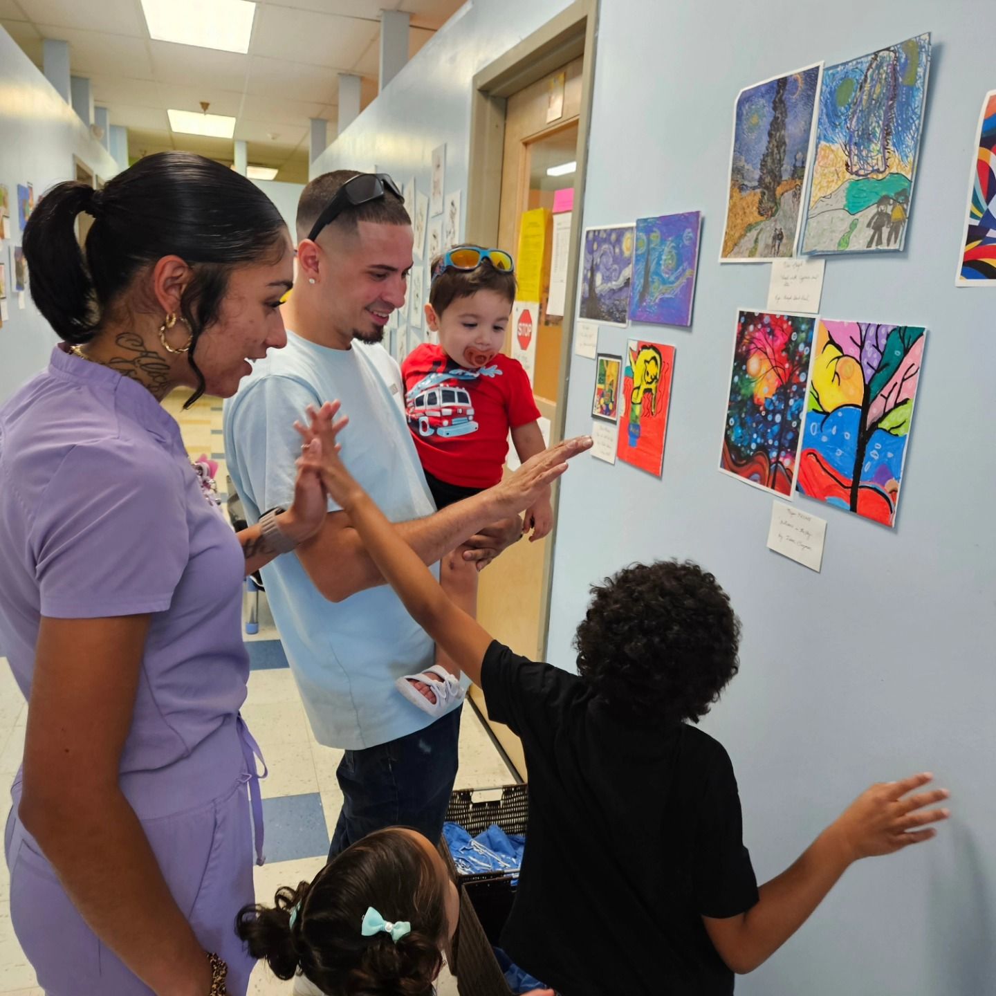 A family looks at colorful children’s artwork displayed on a light blue hallway wall.