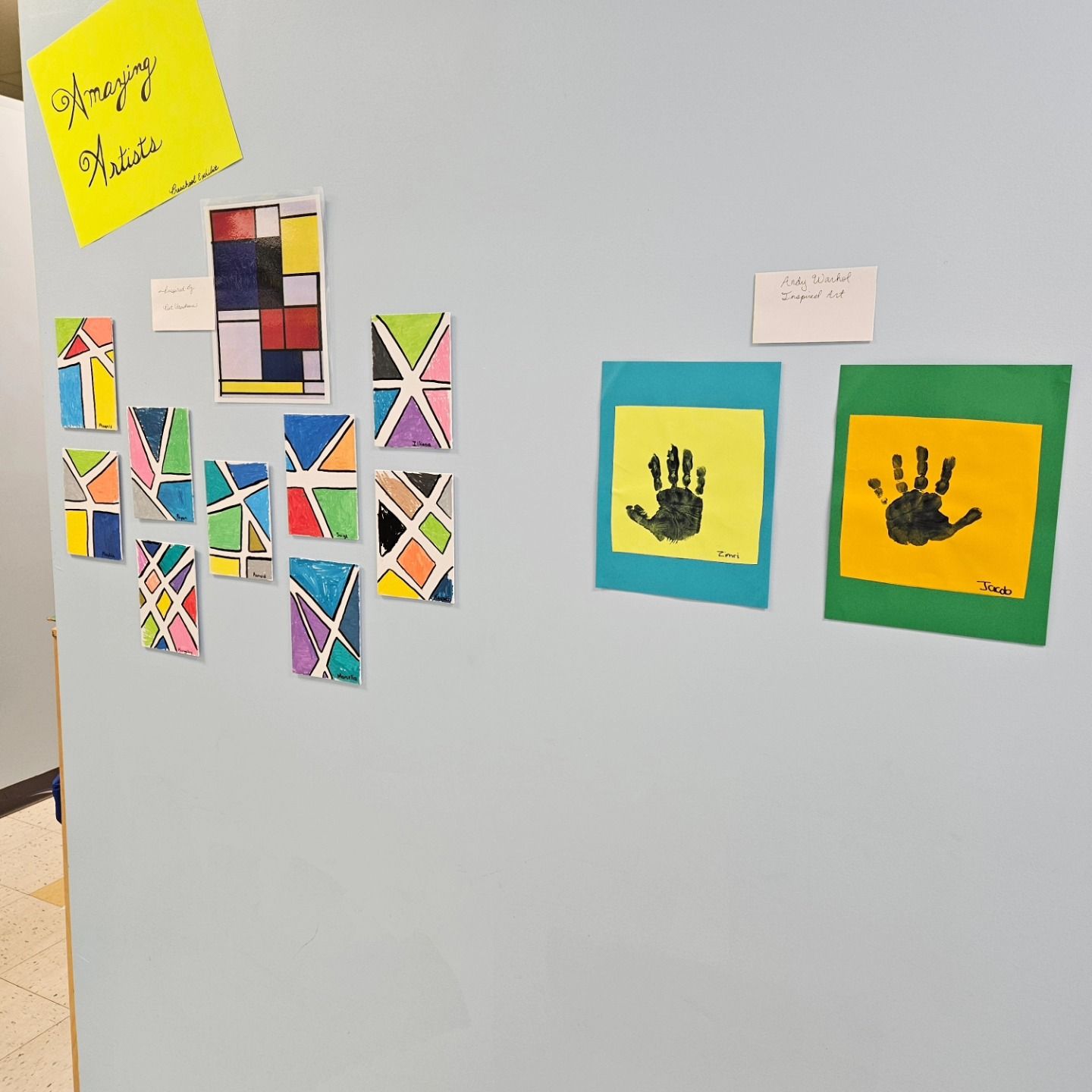 A display of children's art on a wall featuring geometric patterns and two handprint paintings on colored paper.