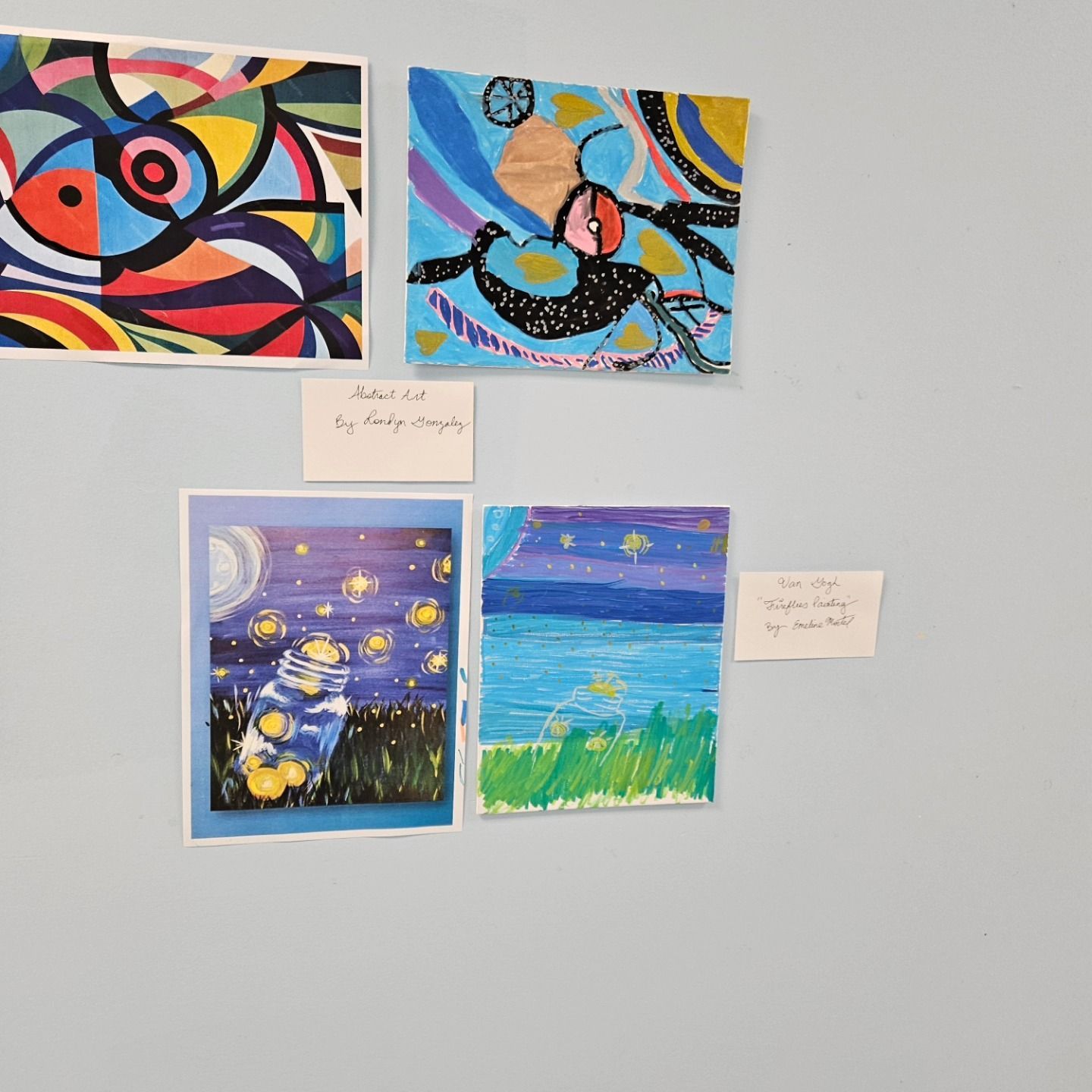 Four colorful paintings are displayed on a light blue wall, each accompanied by a small white note.