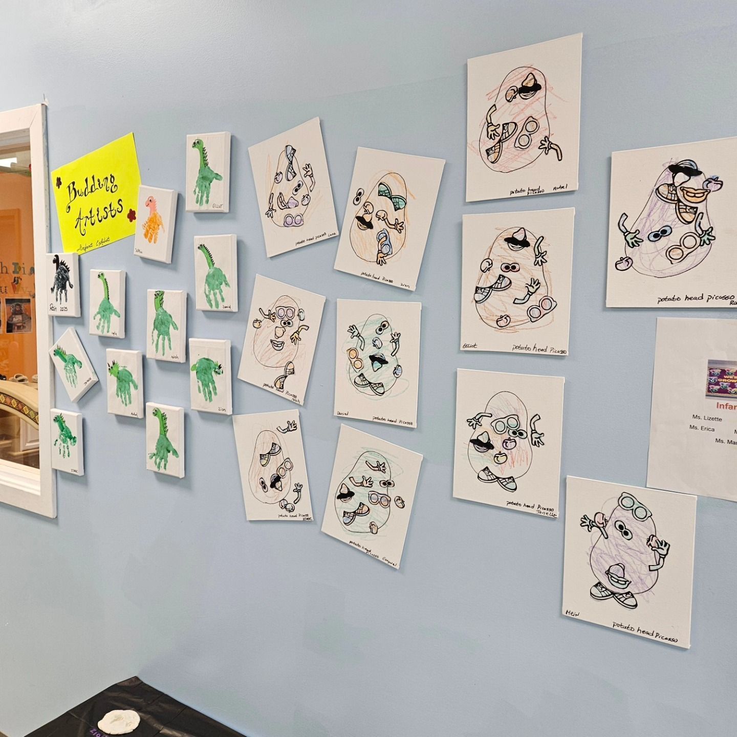 A wall in a classroom displays artwork featuring green handprint giraffes and Mr. Potato Head drawings on white paper.