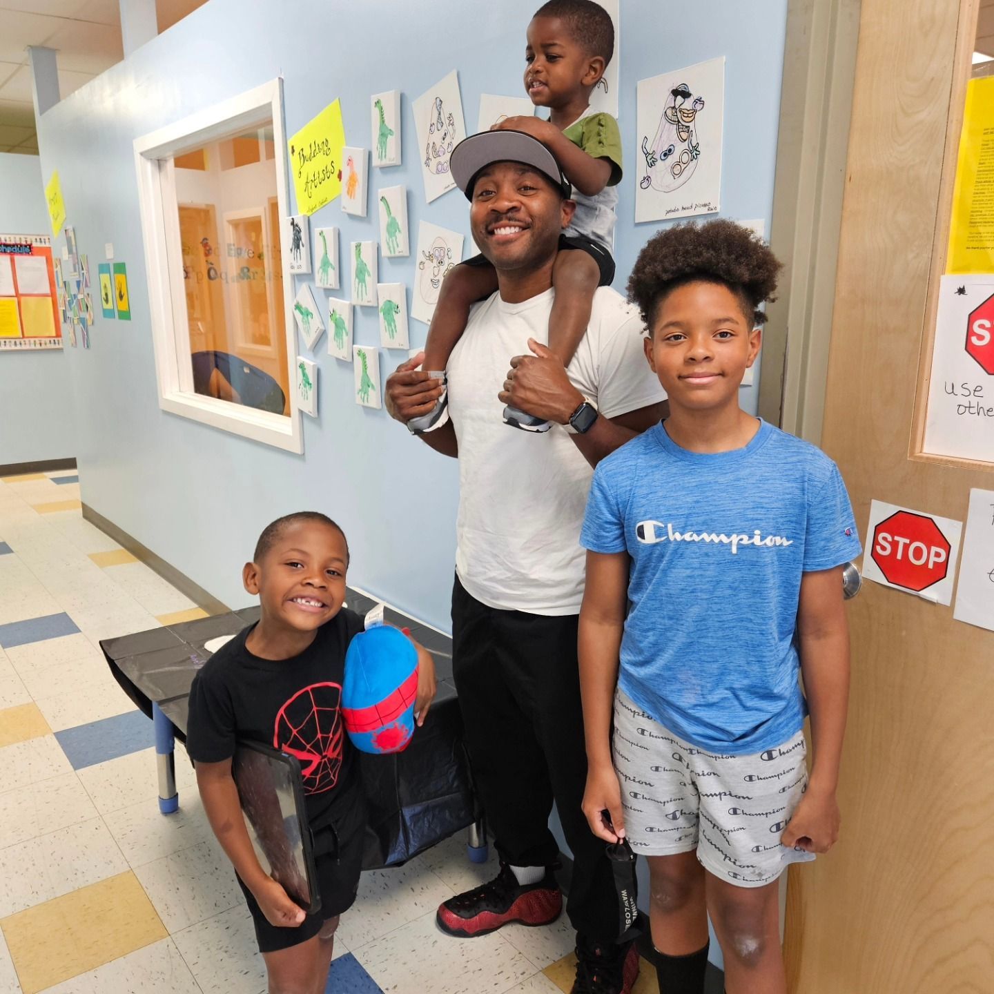 A person stands in a hallway carrying a child on their shoulders, flanked by two others, all smiling near a stop sign.