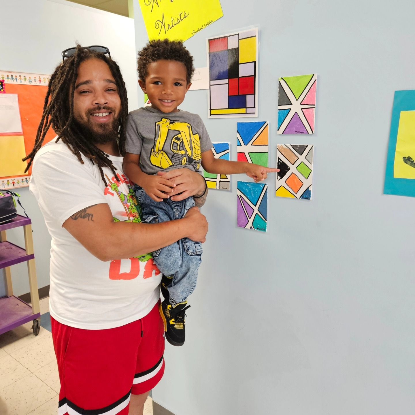 A person holds a child who is pointing at a piece of colorful geometric artwork on a light blue classroom wall.