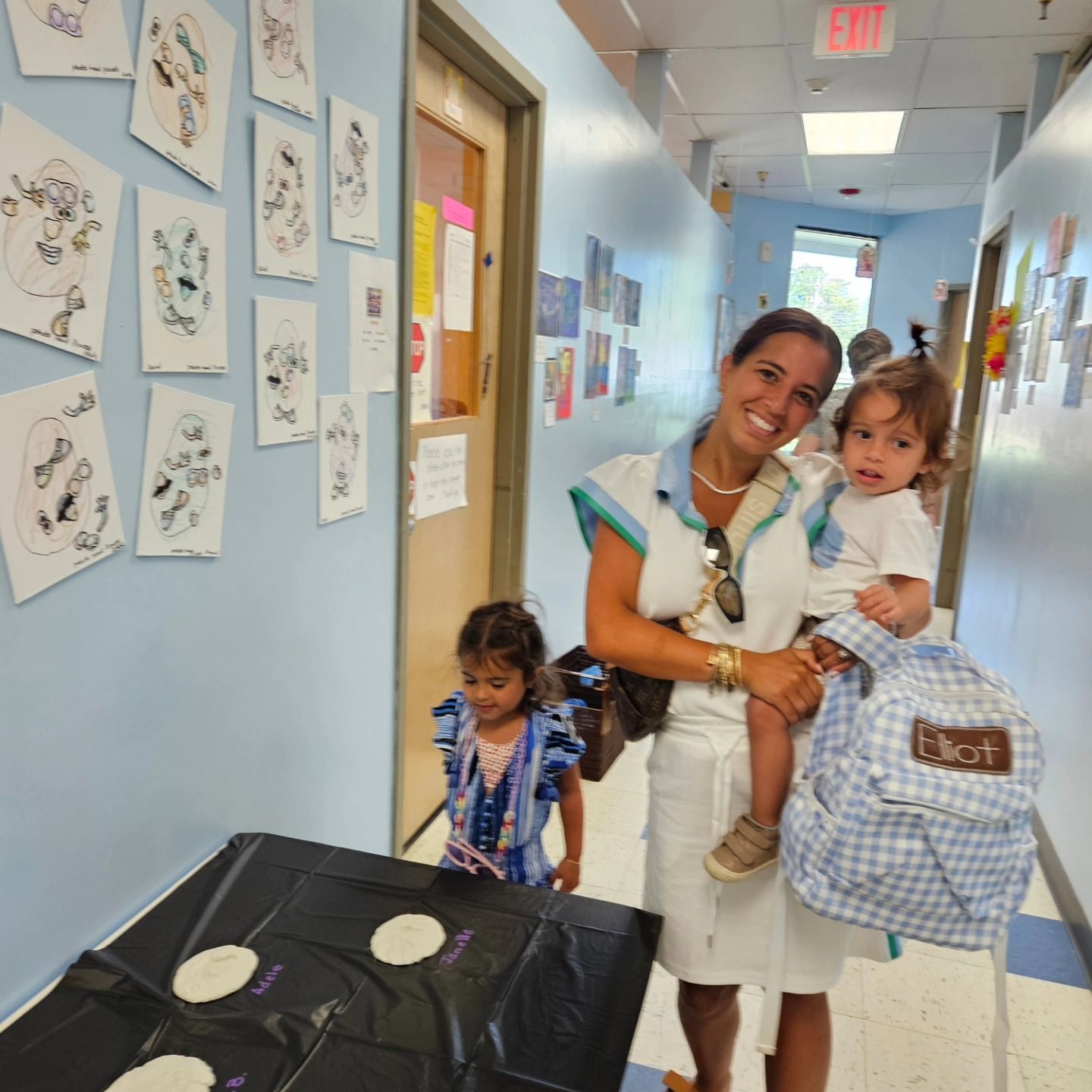 A person carries a child in a school hallway with art displayed on the walls; another child stands nearby.