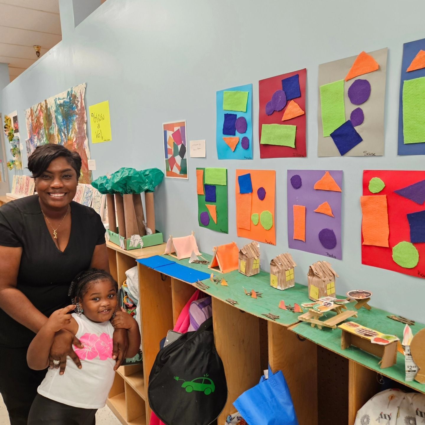 An adult and a child standing in a classroom next to shelves displaying student crafts and art projects on the wall.