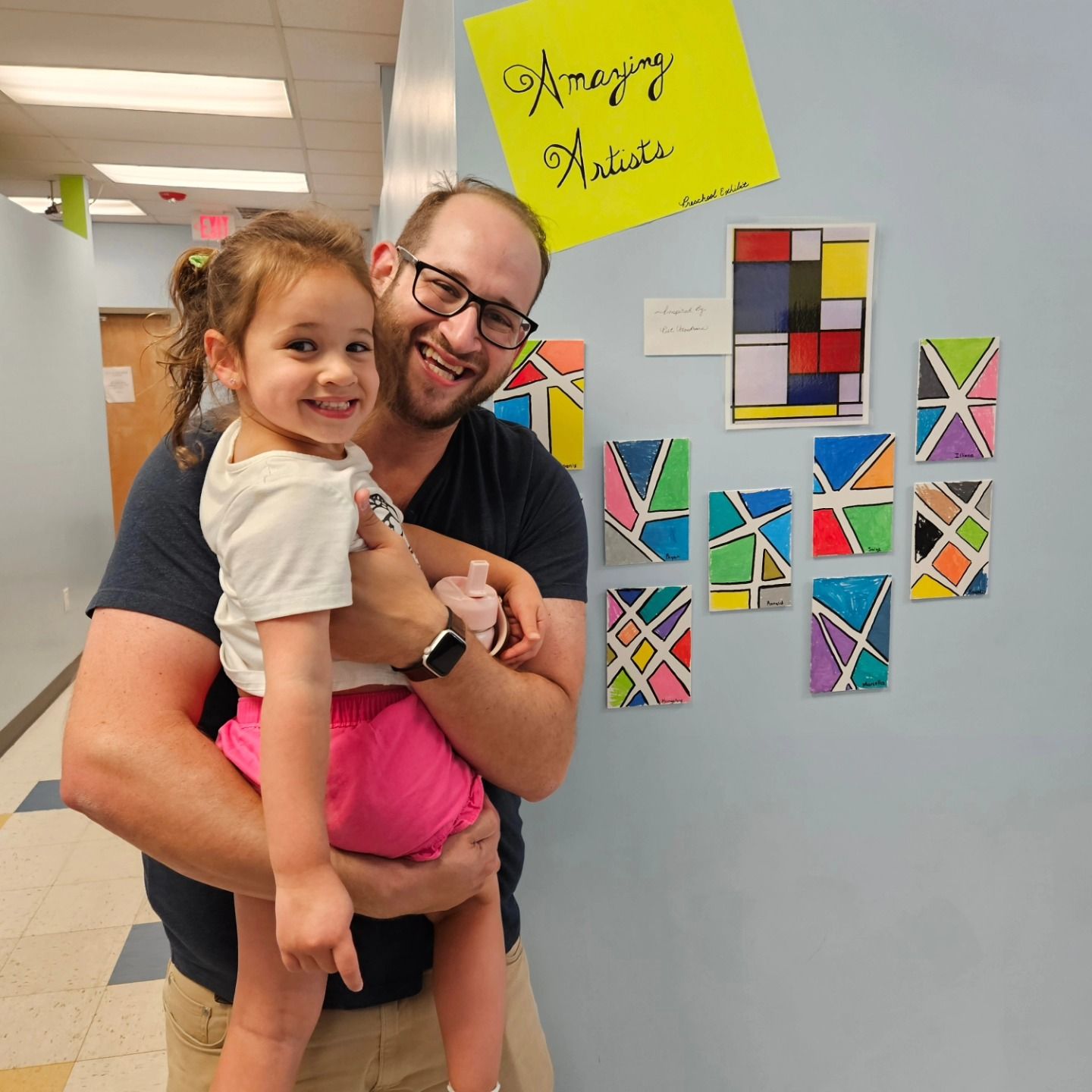 A smiling man holding a child in front of a wall displaying geometric, multicolored children's art.