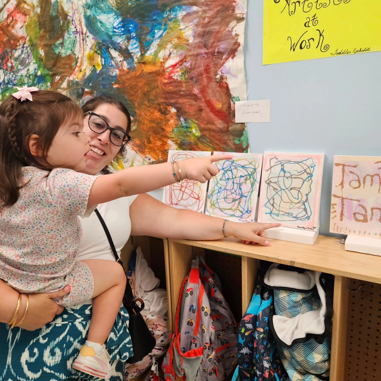 A person holds a child who points at framed colorful abstract drawings on a wooden shelf in front of a painted wall.