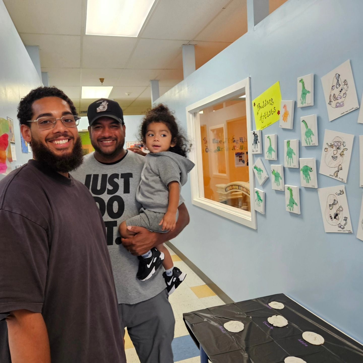 Two people stand in a hallway, one holding a young child. Walls feature hung artwork and a window looks into a classroom.