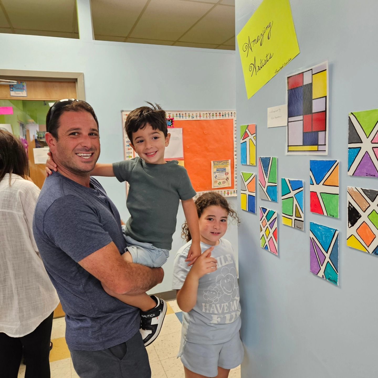 A person holds a child while standing next to another child, both smiling in front of a wall with colorful art projects.