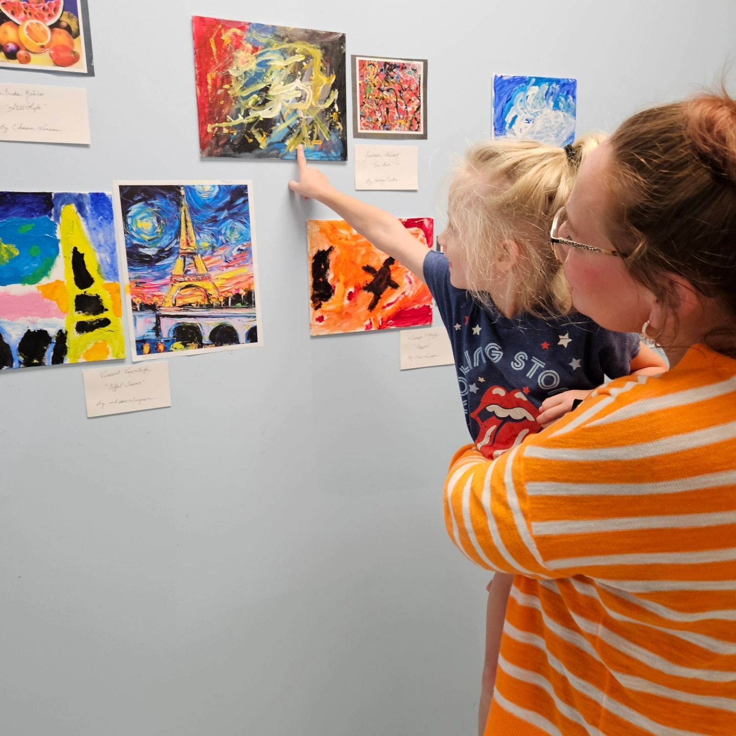 A parent holding a child who points to a colorful, abstract painting displayed on a light blue wall with other artworks.