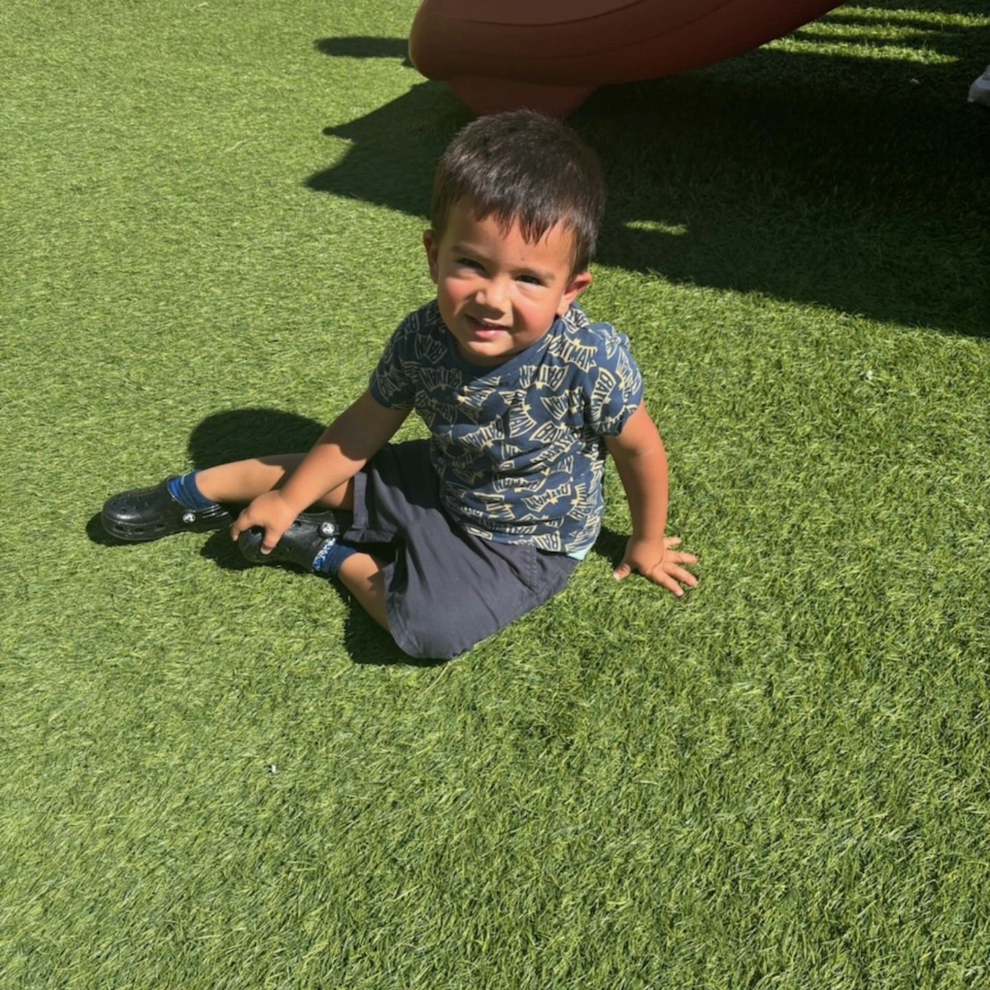 A young child smiling while sitting on bright green artificial grass near a playground structure.