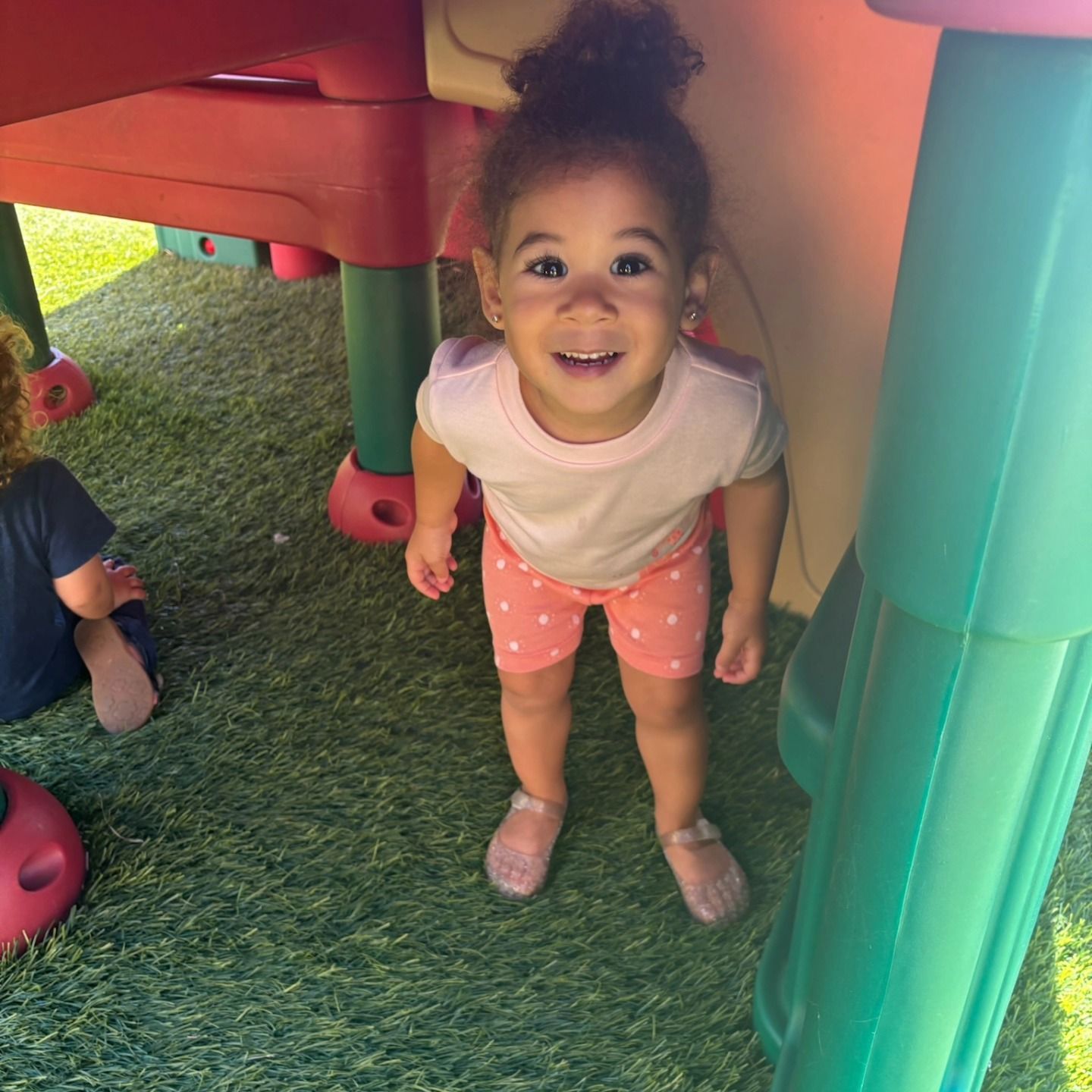 A smiling toddler in a pink shirt and polka-dot shorts stands under a colorful play structure on artificial grass.