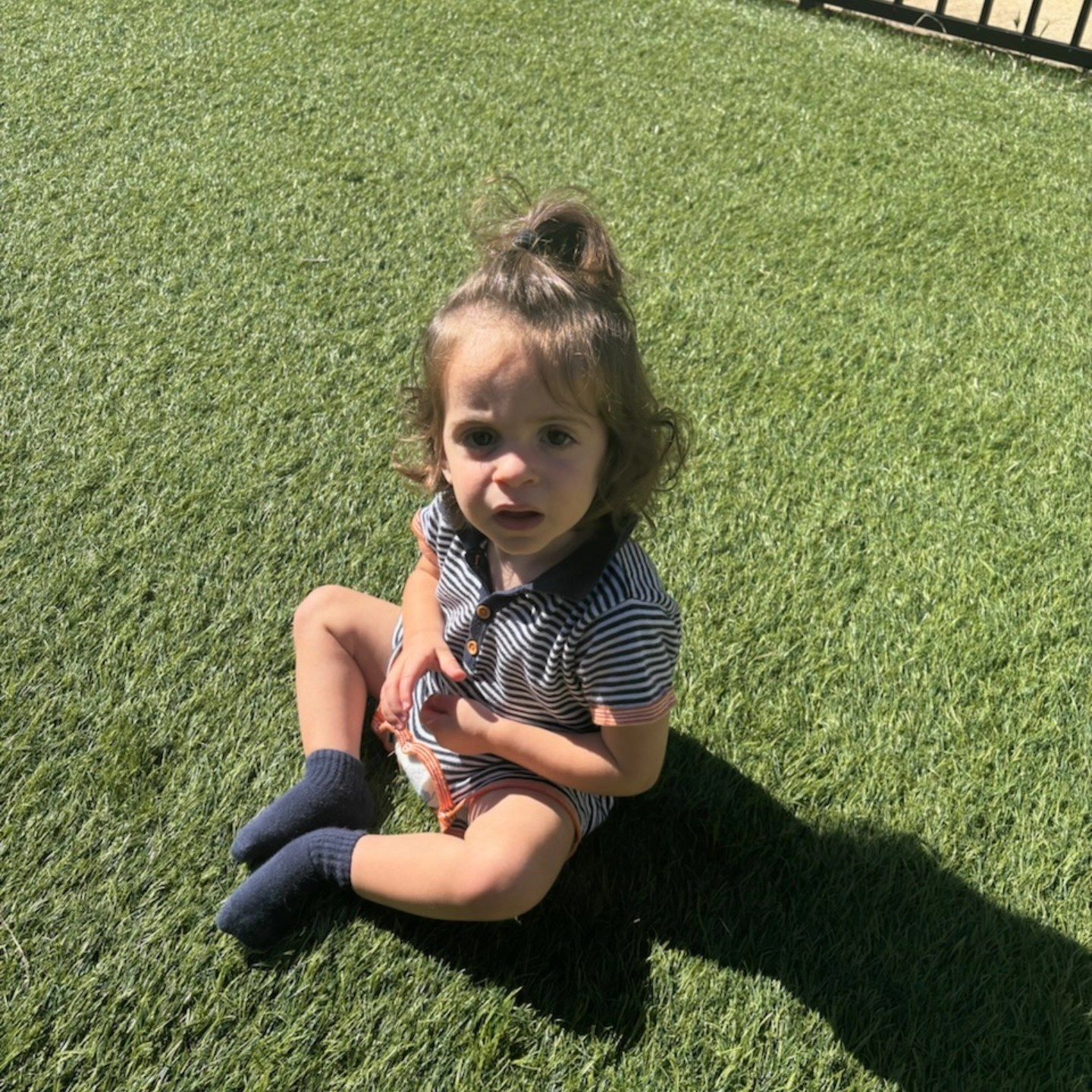 A toddler in a striped polo shirt and blue socks sits on a patch of artificial grass, looking toward the camera.