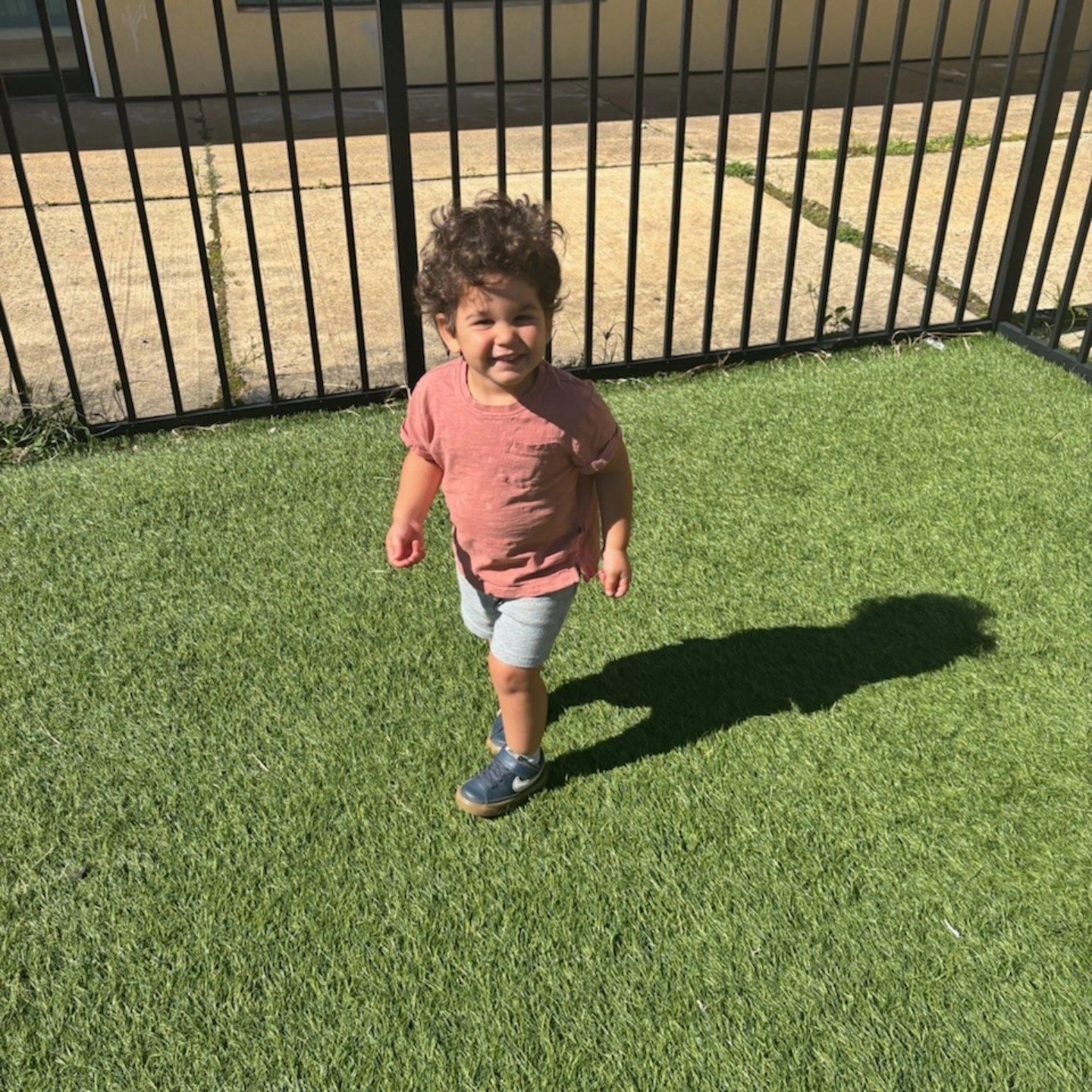 A toddler in a pink shirt and grey shorts stands on artificial grass in front of a black metal fence.