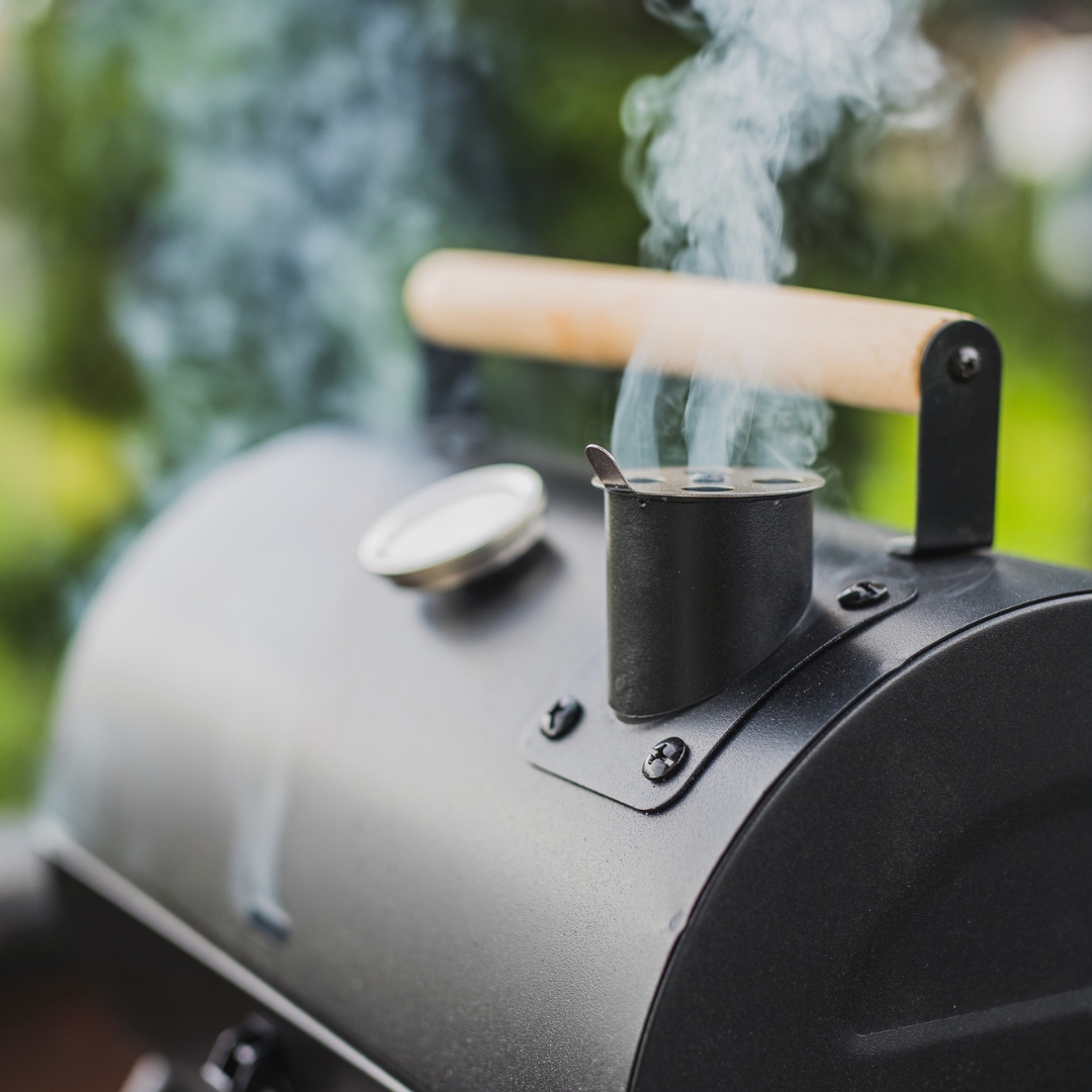 Black smoker grill with wooden handle, releasing smoke outdoors.