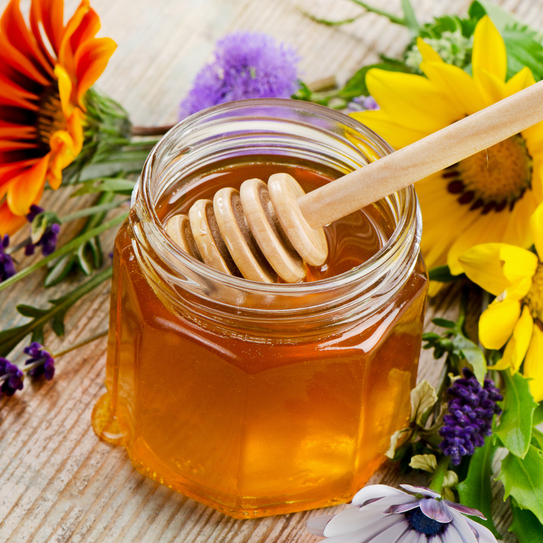 Jar of honey with a wooden dipper surrounded by flowers.