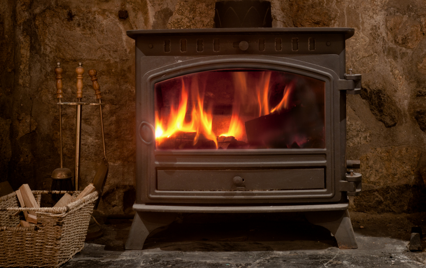 Fire burning brightly inside a dark metal stove set against a stone wall; basket of wood and fireplace tools nearby.