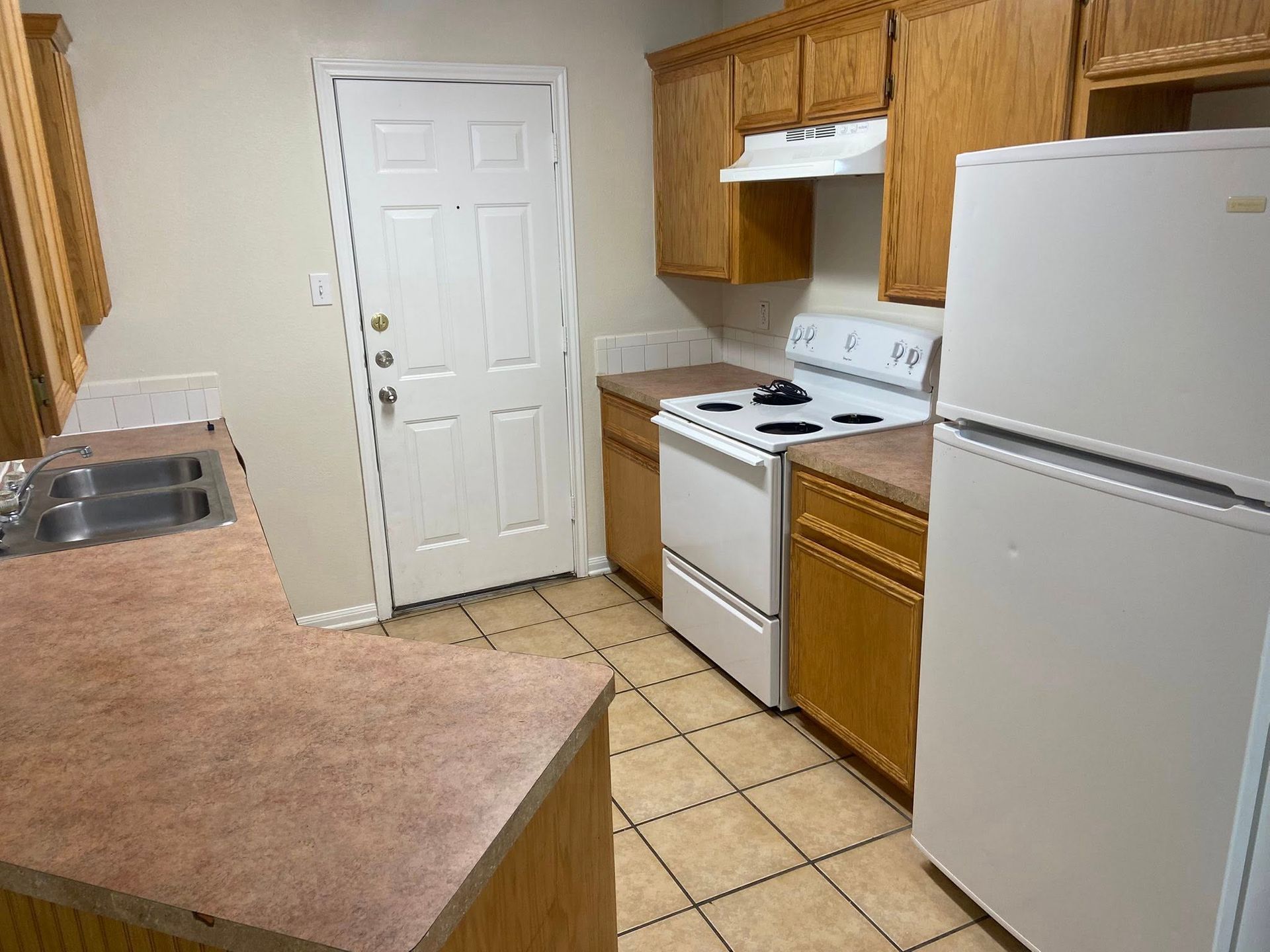 Kitchen with wooden cabinets, white appliances, and a door. Brown countertops and tile floor.