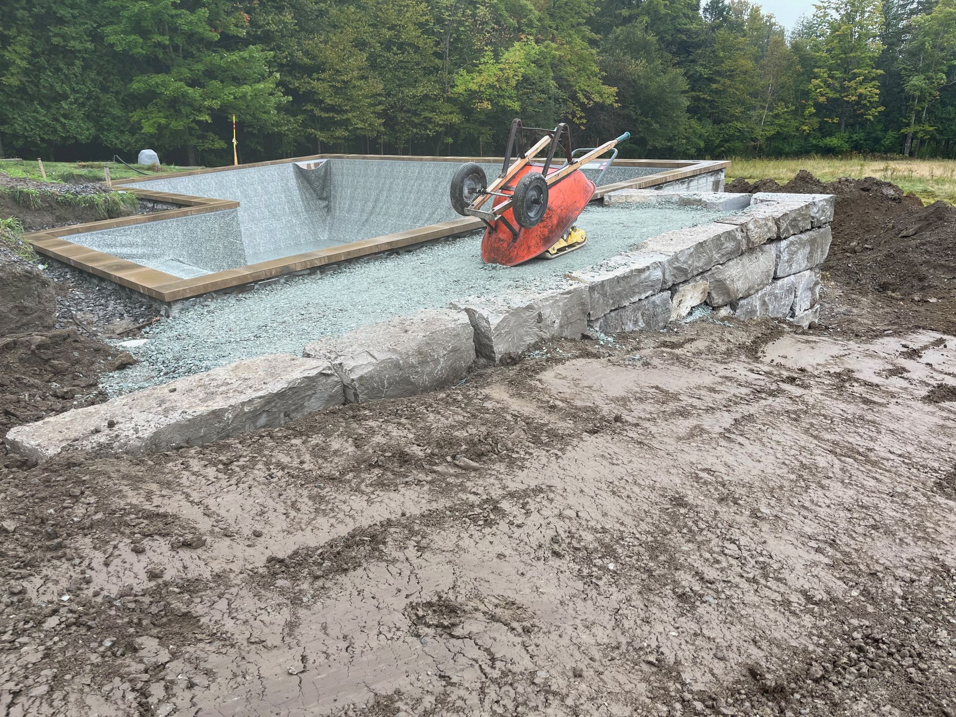 Une brouette est posée sur un tas de terre à côté d'un mur de pierre.