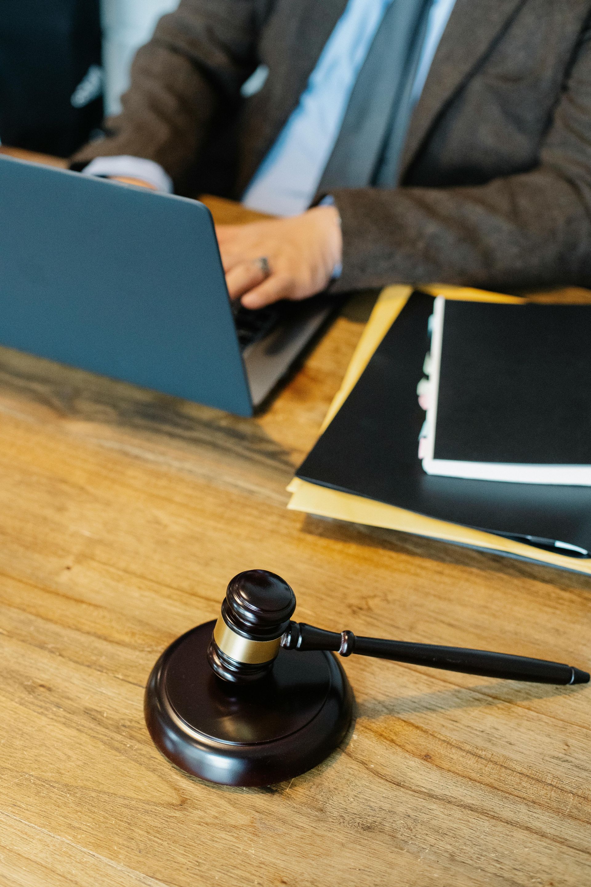 Gavel on wooden table, lawyer typing on laptop, with files in background.