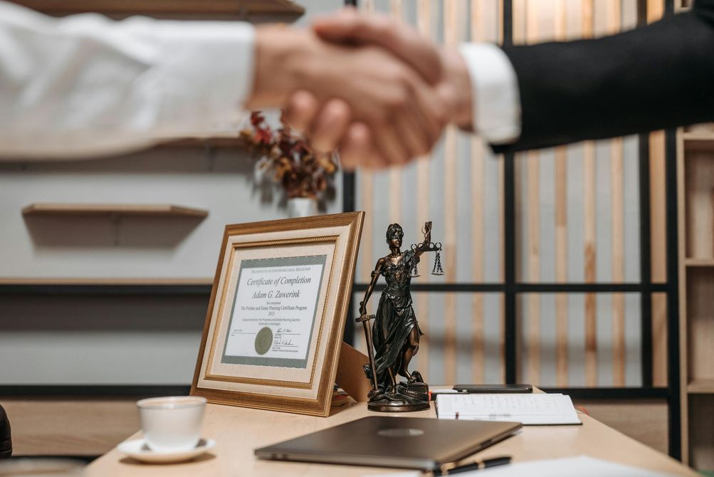 People shaking hands over a desk with a framed certificate and statue of justice.