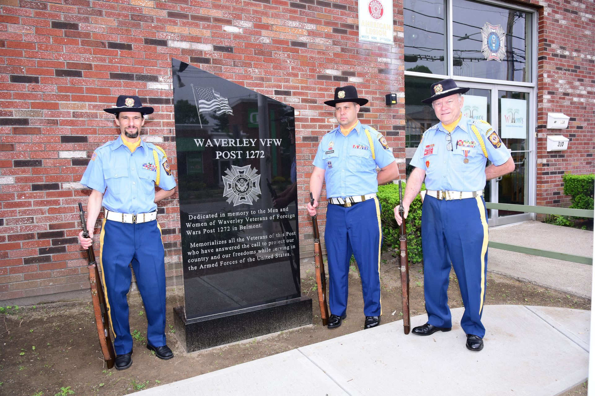 Three men in blue uniforms stand in front of a brick building