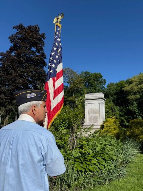 A man in a blue shirt stands in front of an American flag