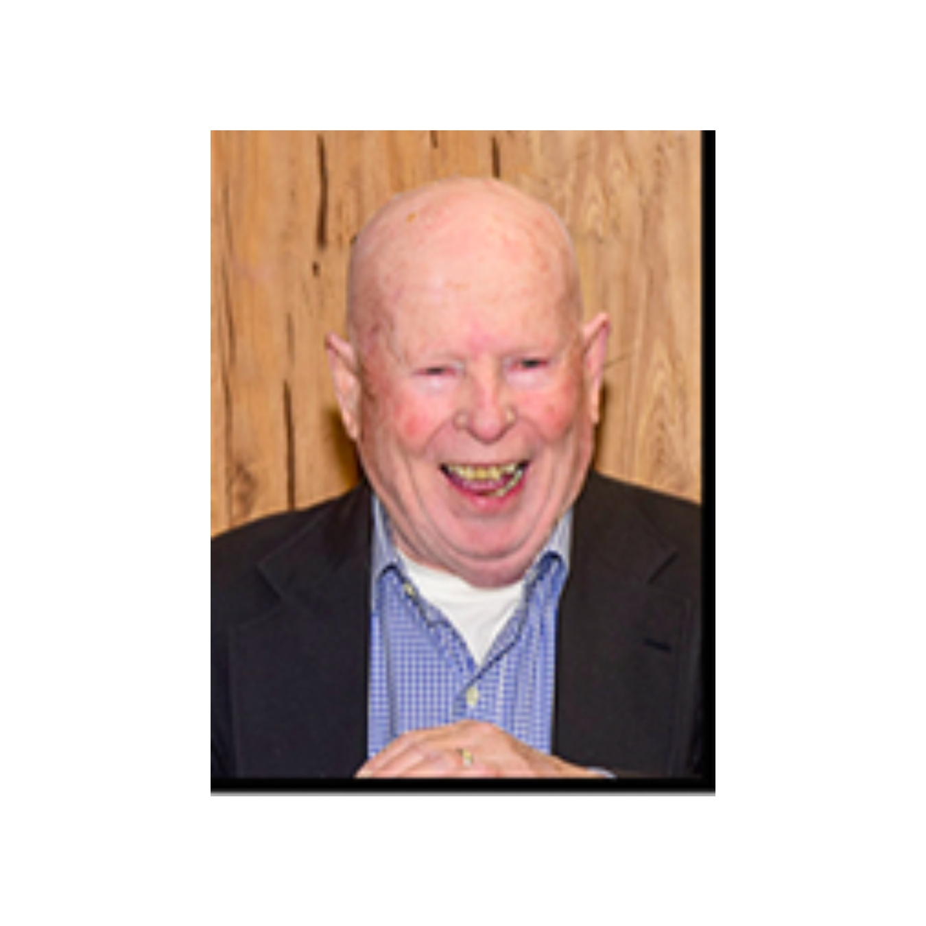 An elderly man in a suit and blue plaid shirt is smiling.