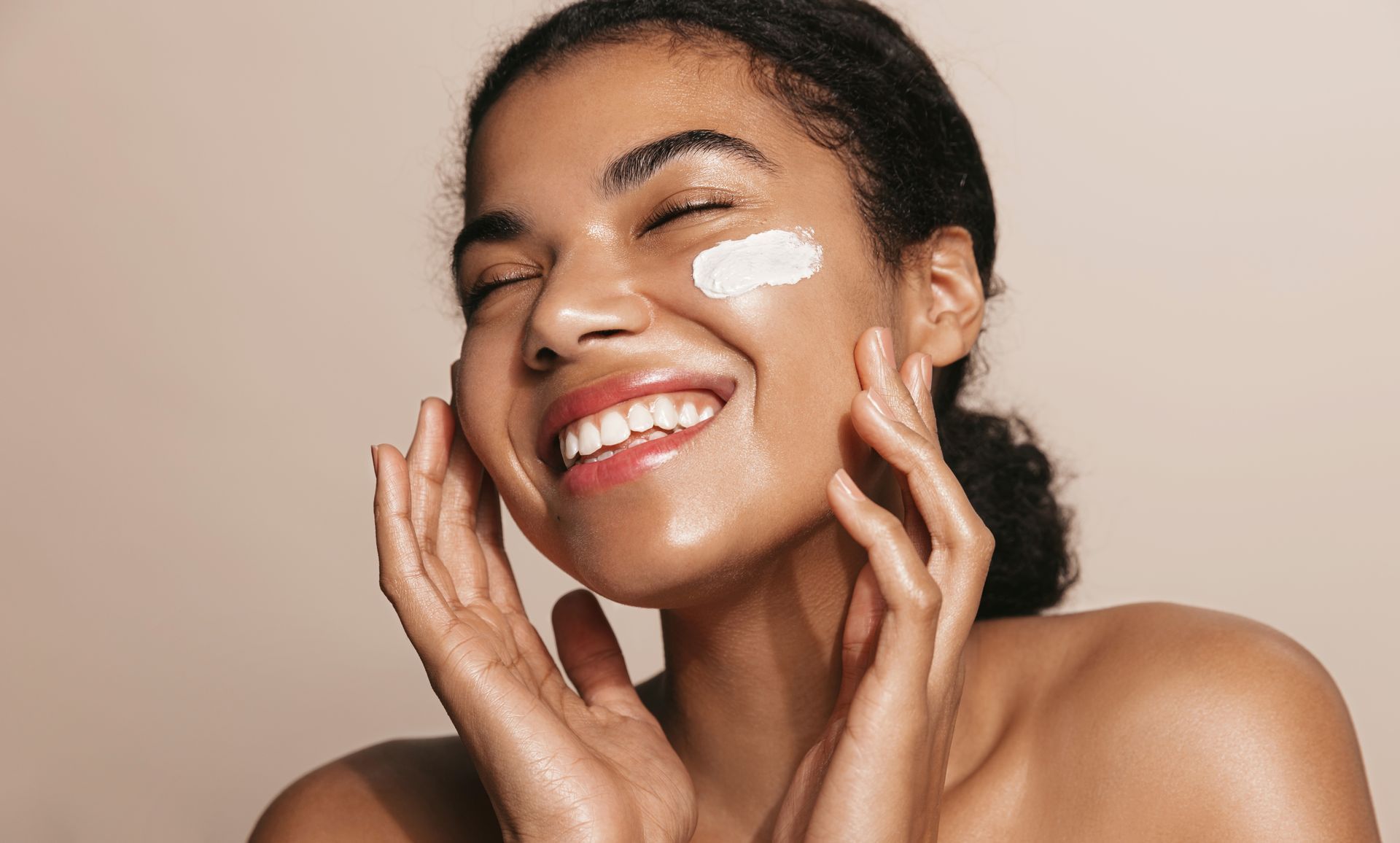 A woman is smiling while applying cream to her face.