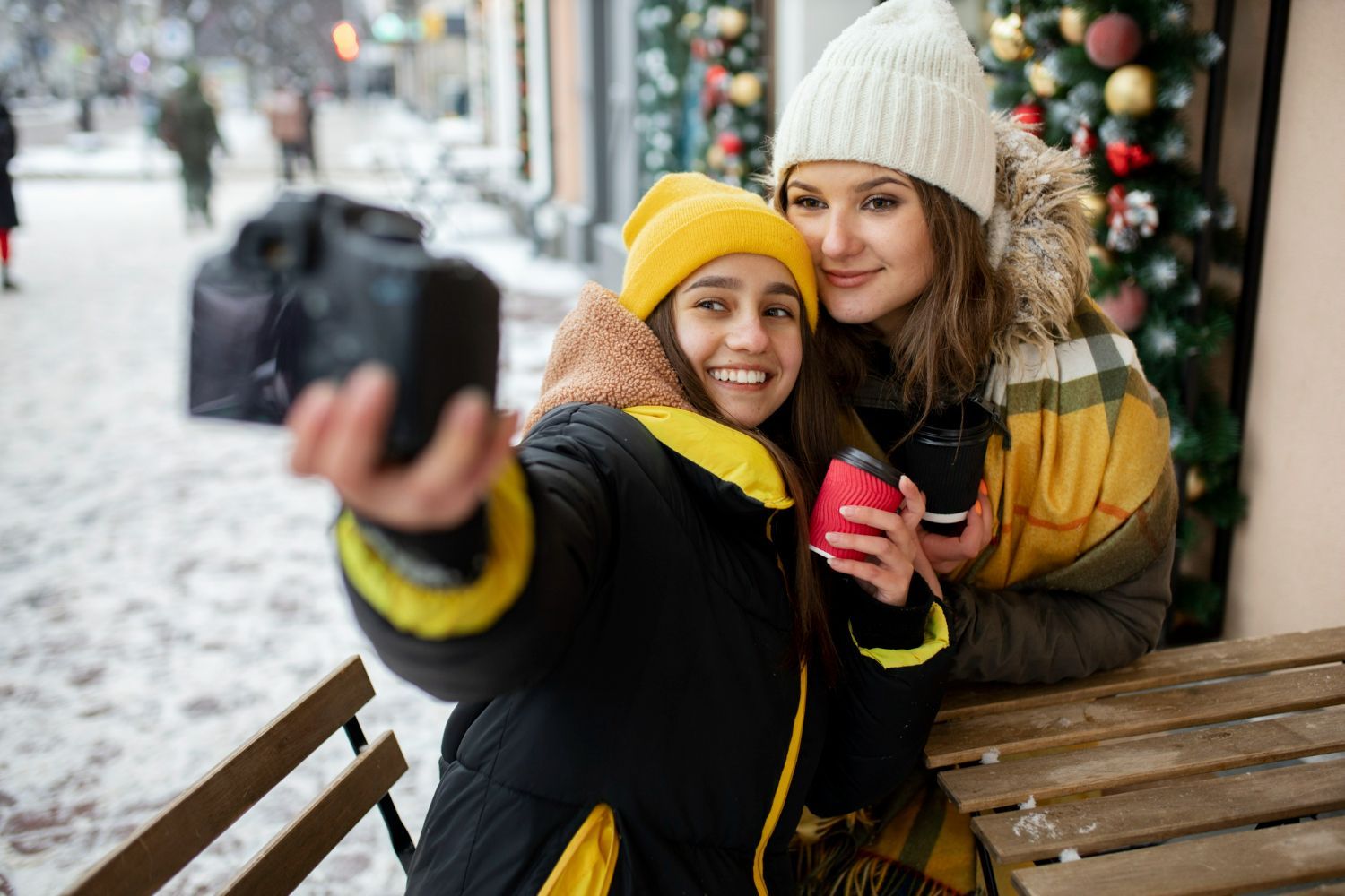 Woman and girl taking a selfie outdoors in winter; girl holding camera, both smiling, with snow and storefront visible.