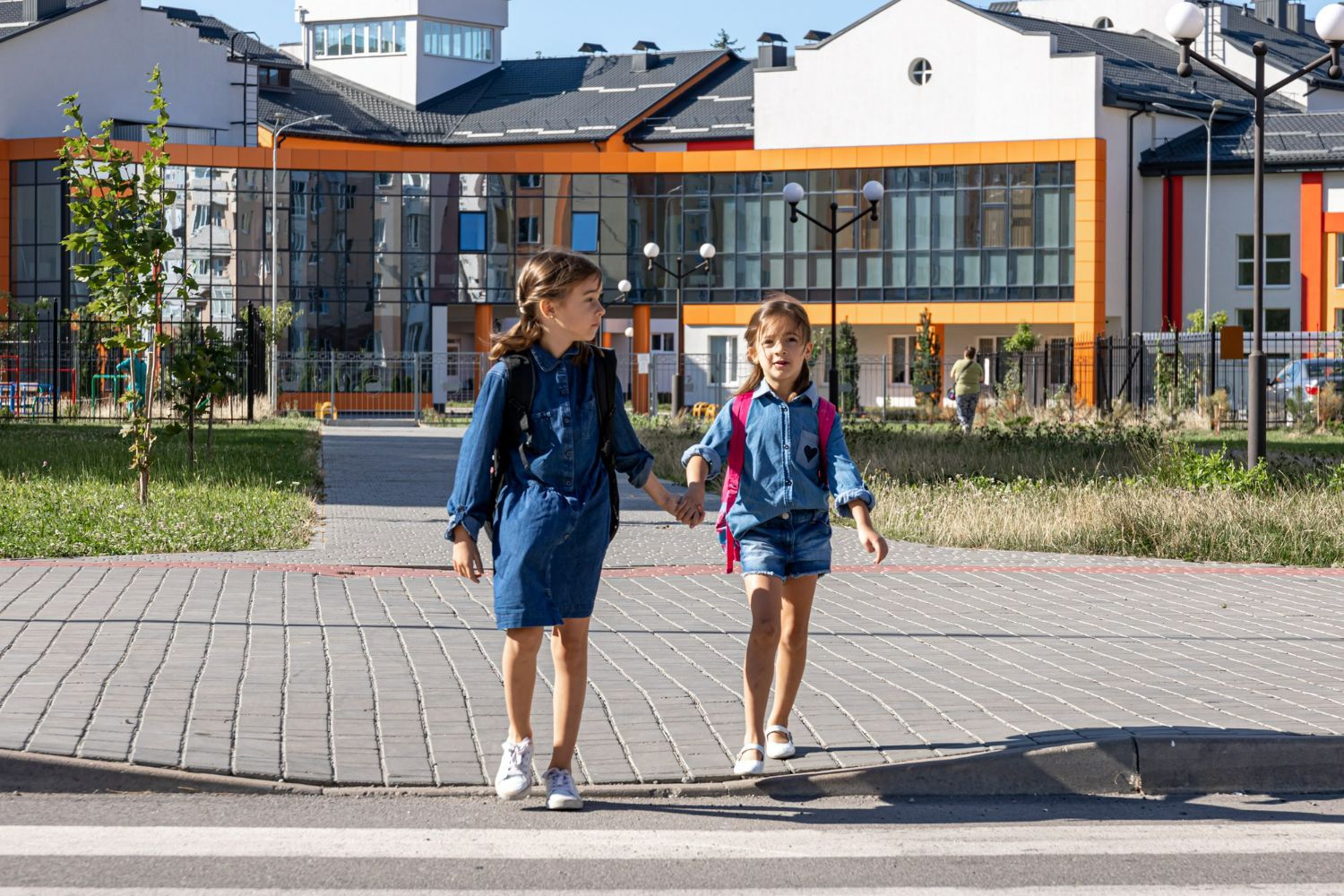 Two little girls are holding hands while crossing the street in front of a school.