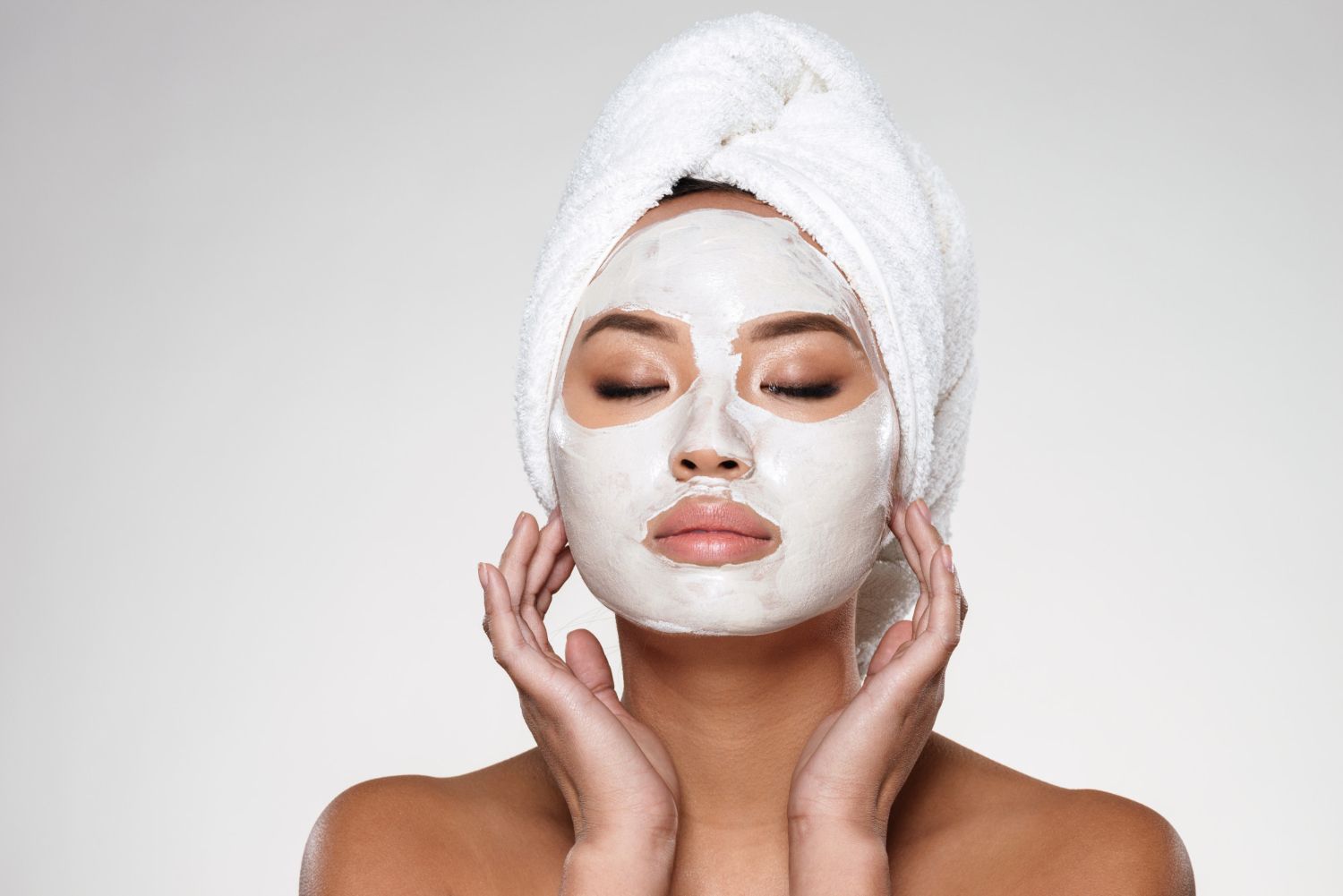 Person with a white facial mask and towel wrapped around head, gently touching their cheeks.