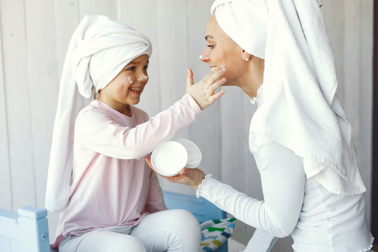 Mother and child with towels on heads, applying cream, smiling. Indoors.