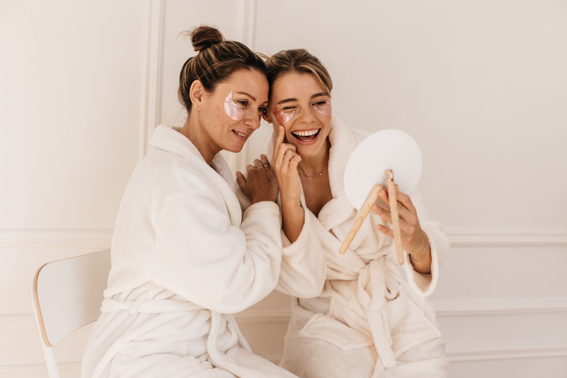 Two women in white robes apply eye patches, one smiles at a mirror. Neutral background.