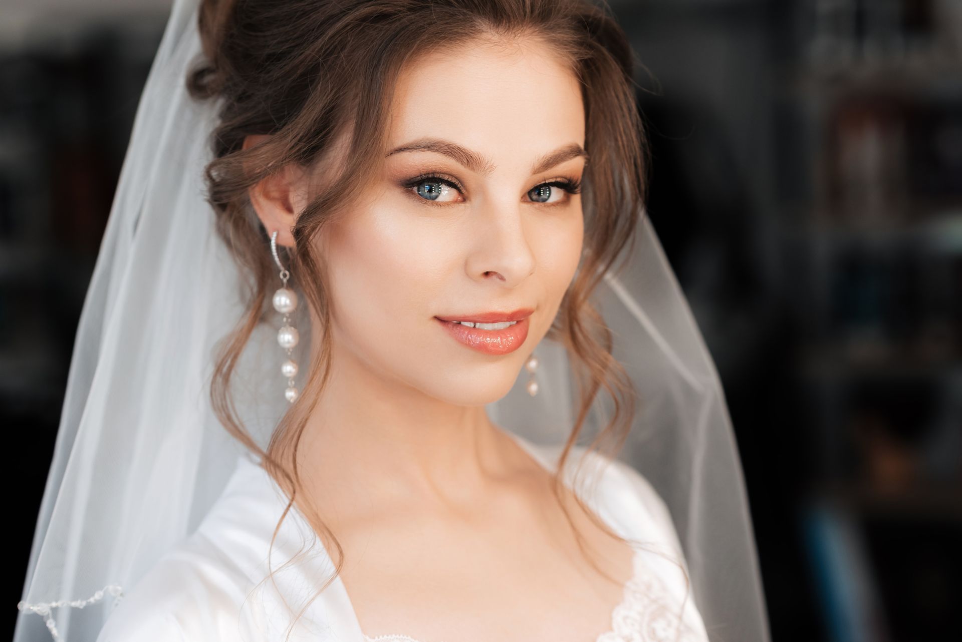 Bride in a white veil and dress smiling softly, with pearl earrings against a blurred dark background