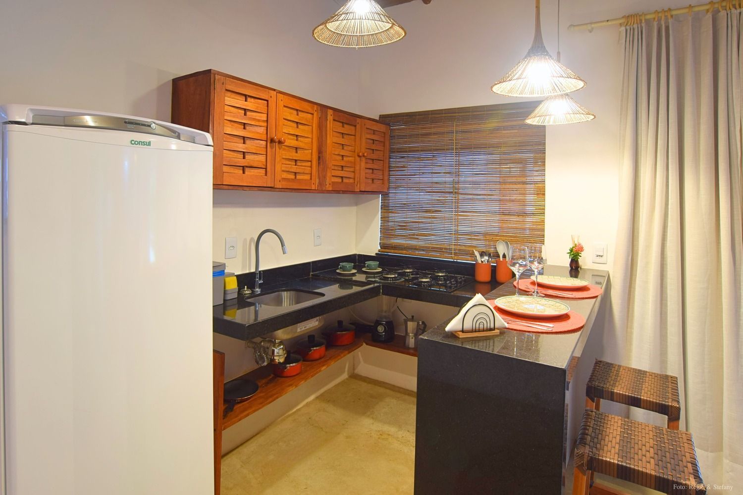 Cozinha pequena com armários de madeira, bancadas pretas e balcão para refeições rápidas. Geladeira branca à esquerda.