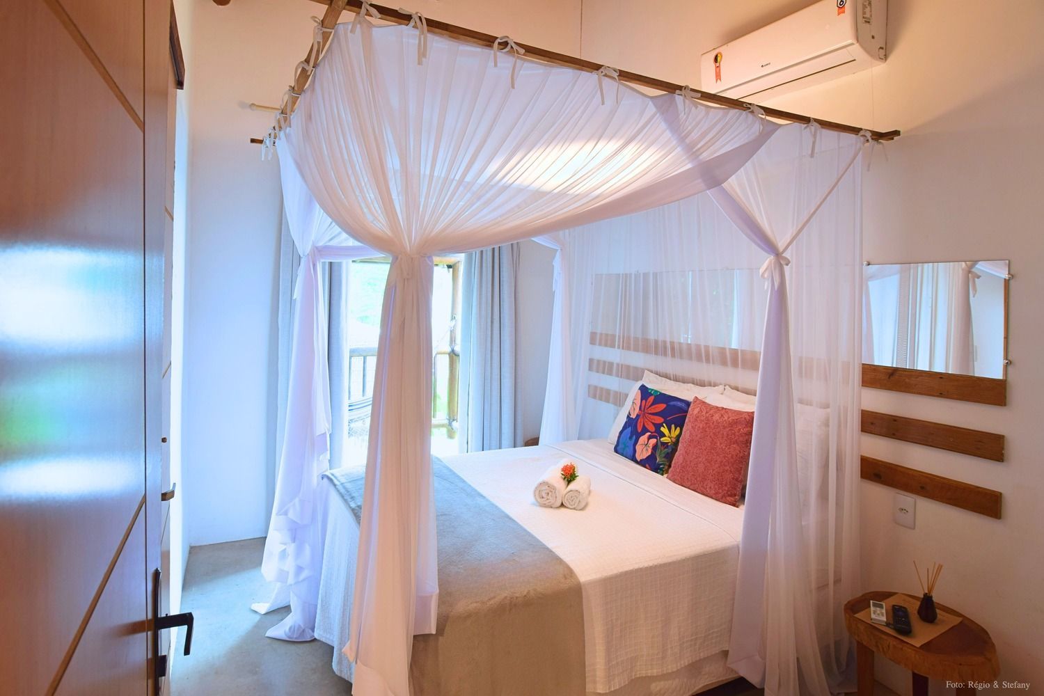 Quarto com cama de dossel, lençóis brancos, almofadas coloridas e acesso à varanda.