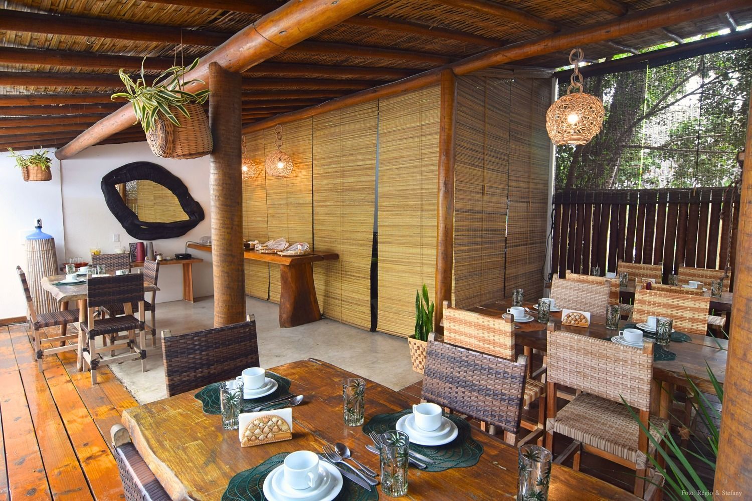Área de jantar ao ar livre com móveis de madeira, detalhes em bambu e plantas suspensas.
