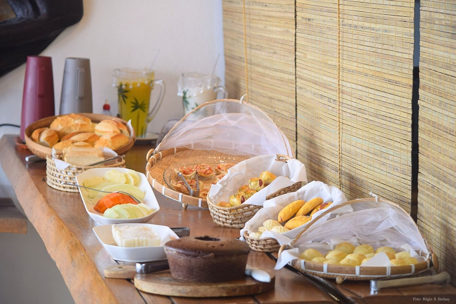 Buffet de café da manhã: pão, doces, bolo, queijo, frutas e suco sobre uma mesa de madeira.
