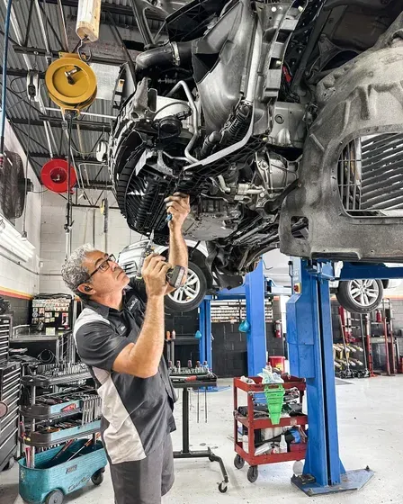 Mechanic works on a car lifted in a shop. He uses a tool under the car, which is black.