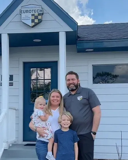 Family in front of Eurotech building. Woman holding a child, boy, and man with beard smiling, all in front of blue door.