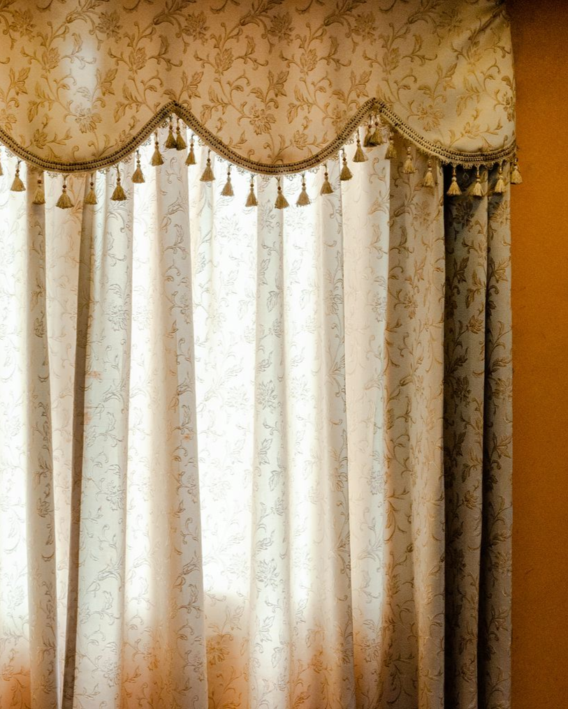 Beige patterned curtains with a scalloped valance and tassel trim hanging over a sunlit window