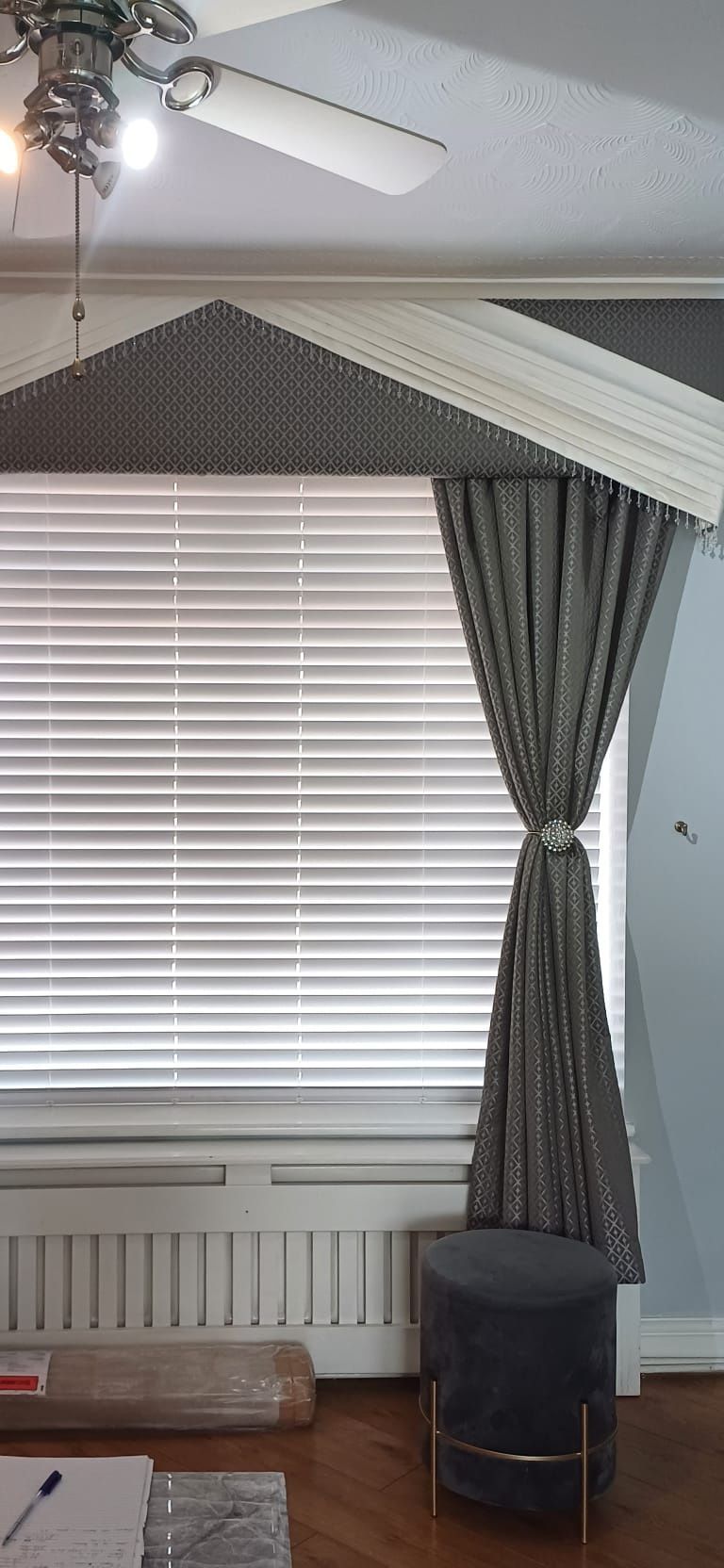 Living room window with white blinds, gray curtains, radiator, and a dark ottoman beneath a peaked ceiling.