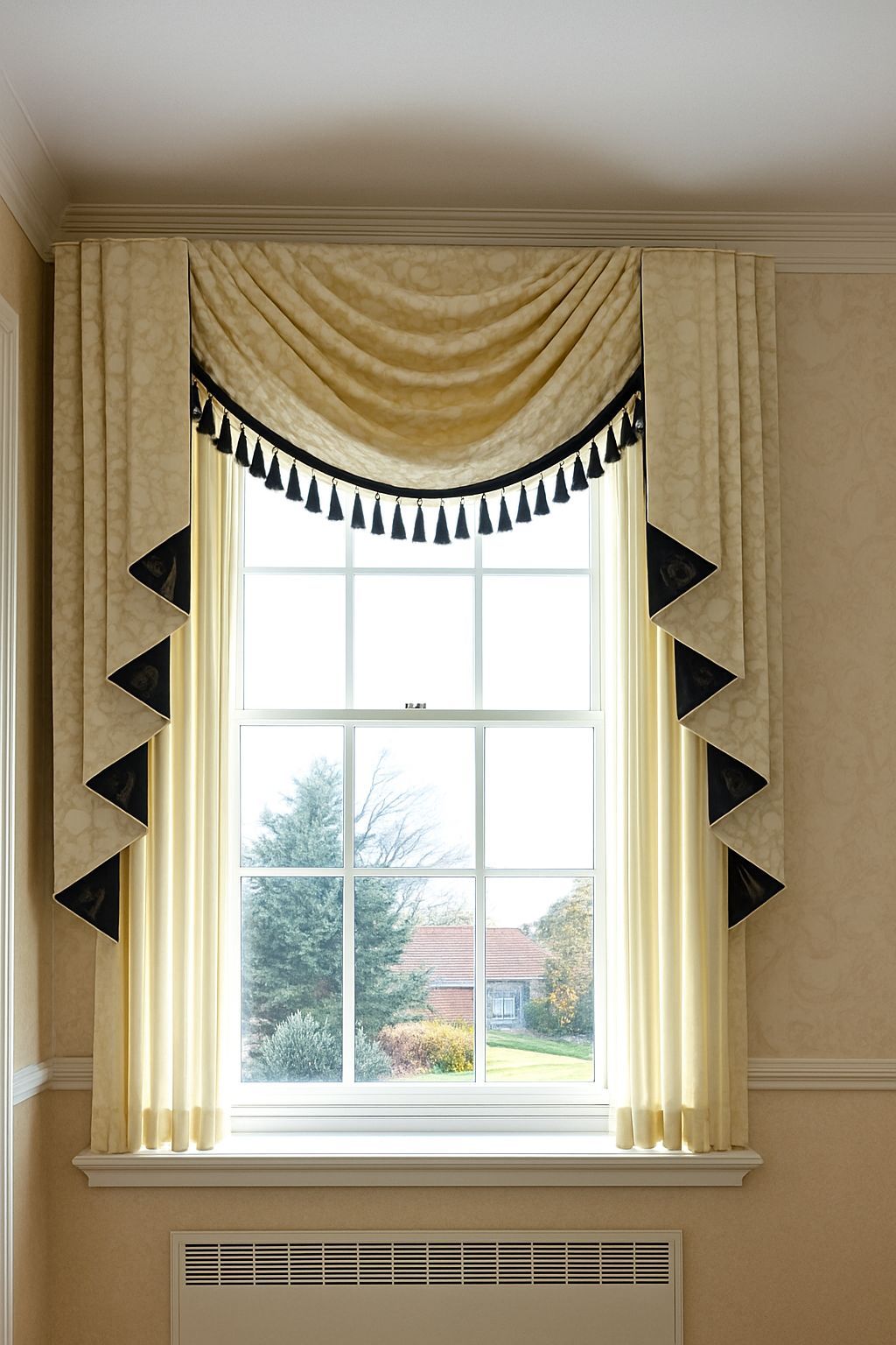 Beige window drapery with a swag valance and black tassel trim in a room.