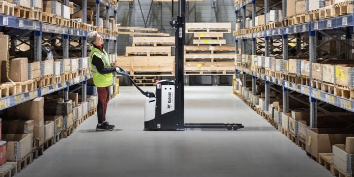 A man is standing next to a stacker in a warehouse.