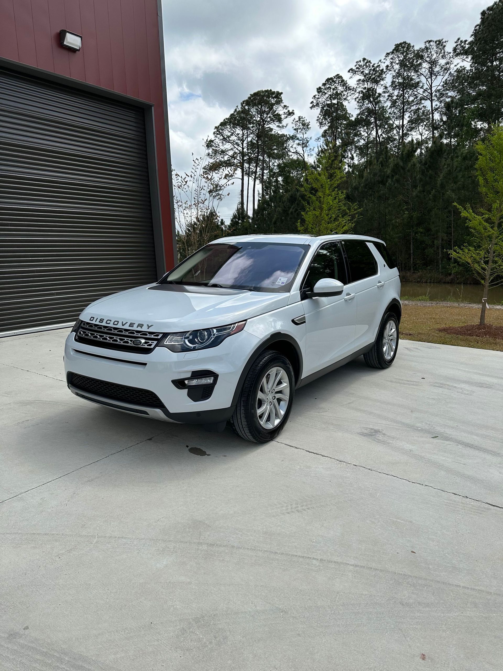 White SUV parked outside a red building on a concrete driveway, with trees in the background.