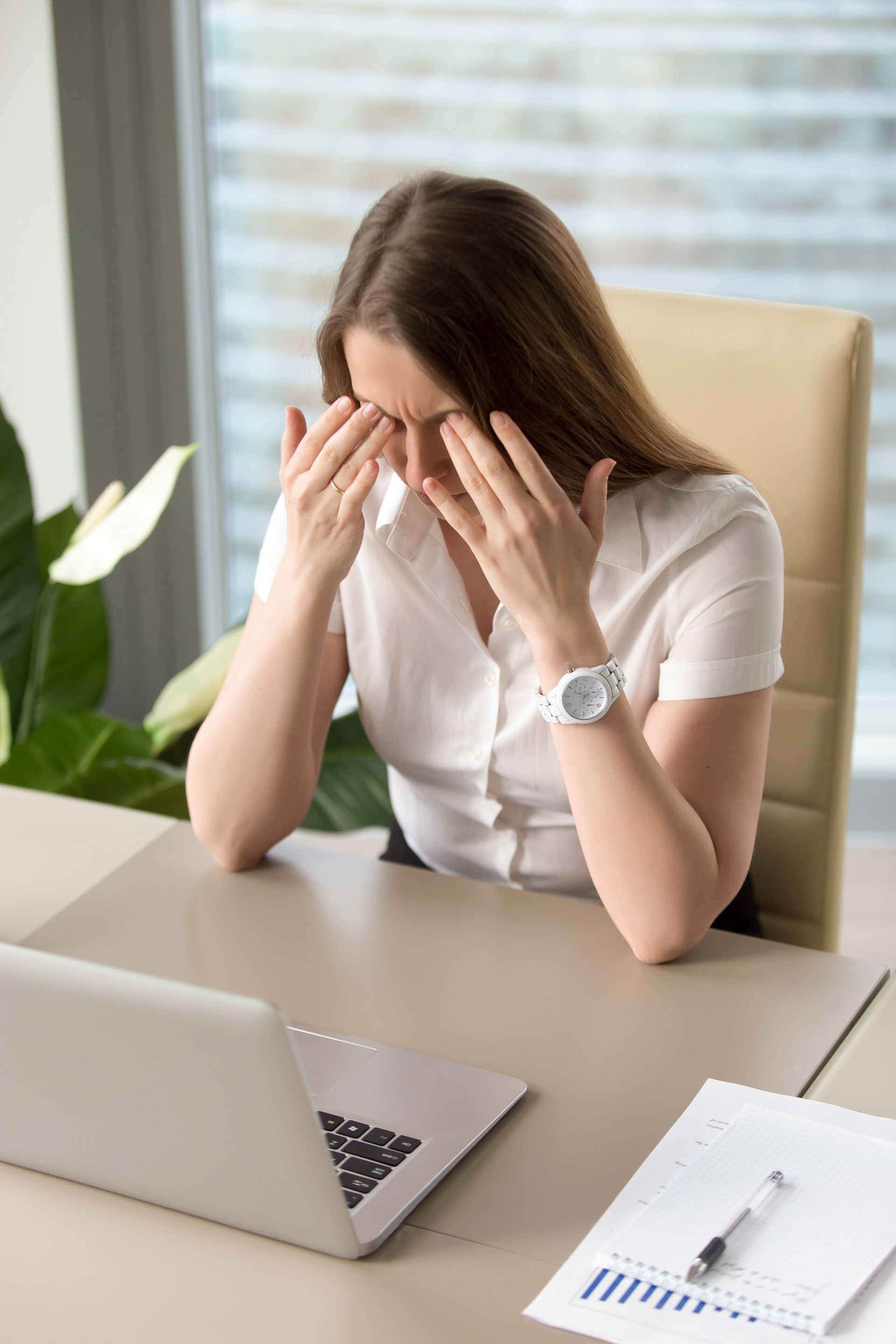 A woman is sitting at a desk with a laptop and blowing her nose.