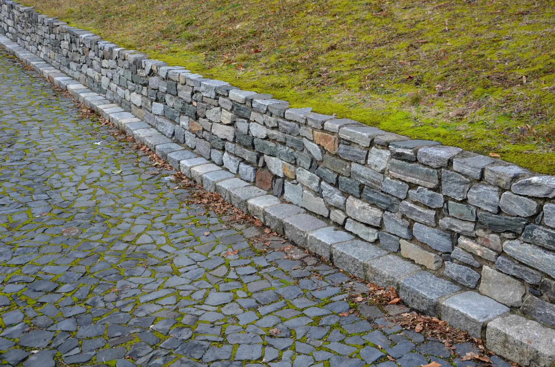 Stone wall bordering cobblestone path and grassy hill.