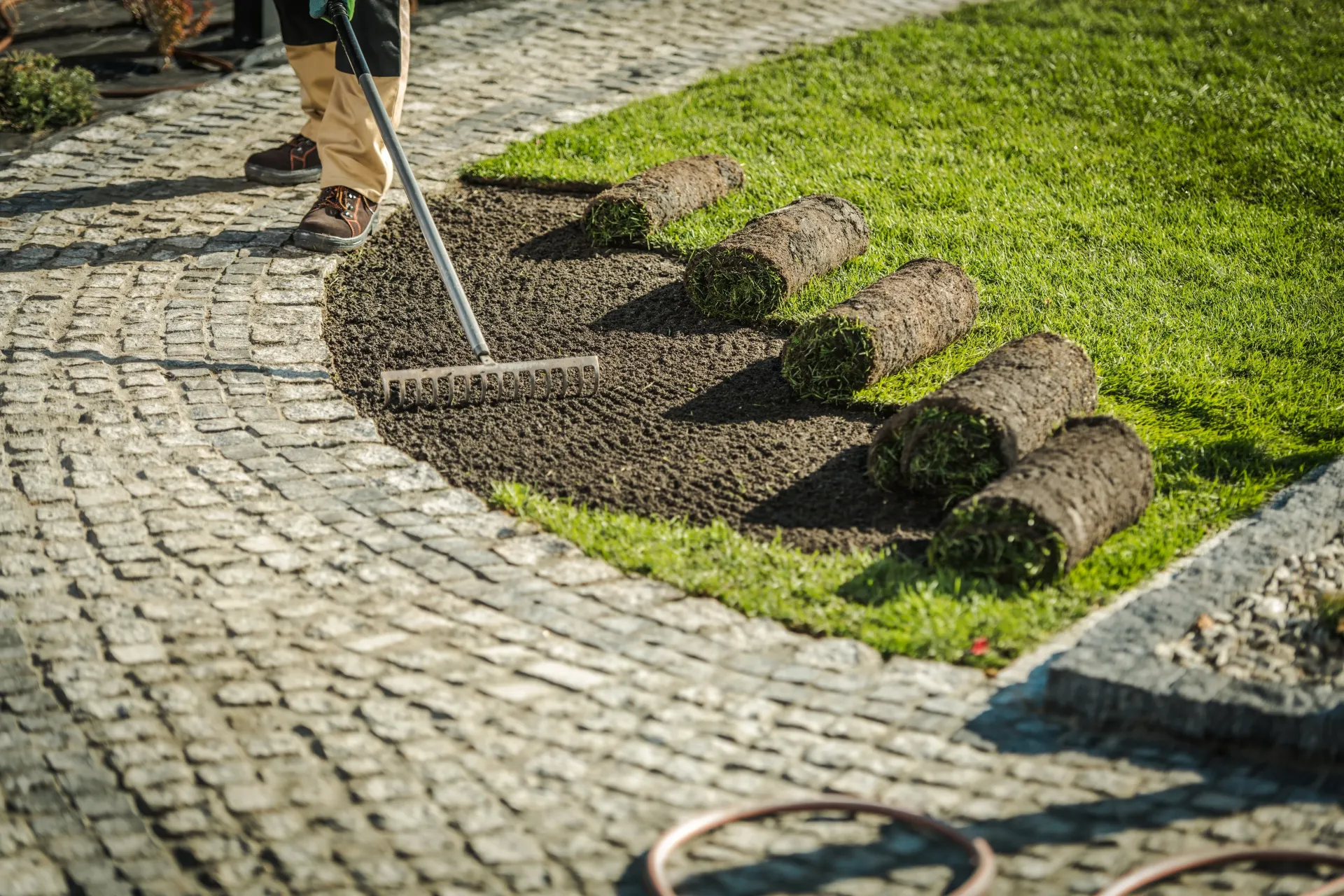 Person raking soil near a stone walkway, preparing to lay rolls of fresh sod onto the ground.