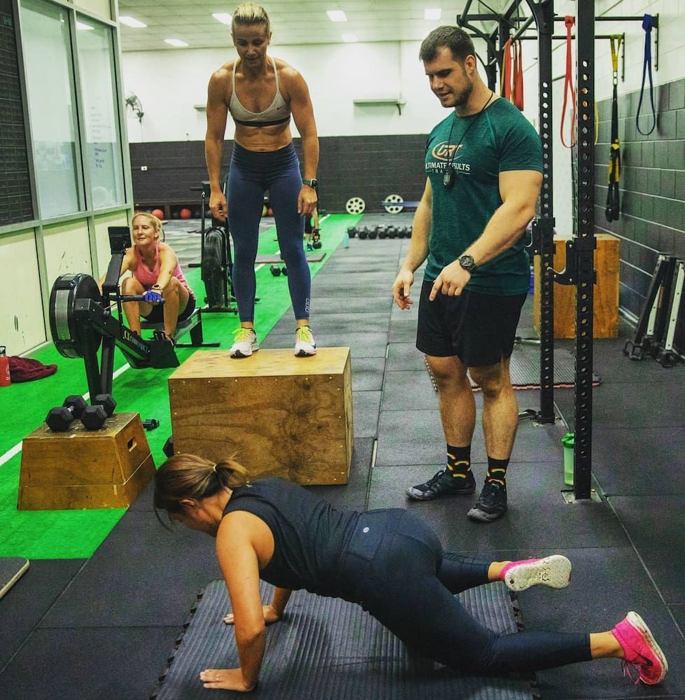 A Man In A Green Shirt Is Standing Next To A Woman Doing Push Ups — Ultimate Results Training in Aitkenvale, QLD