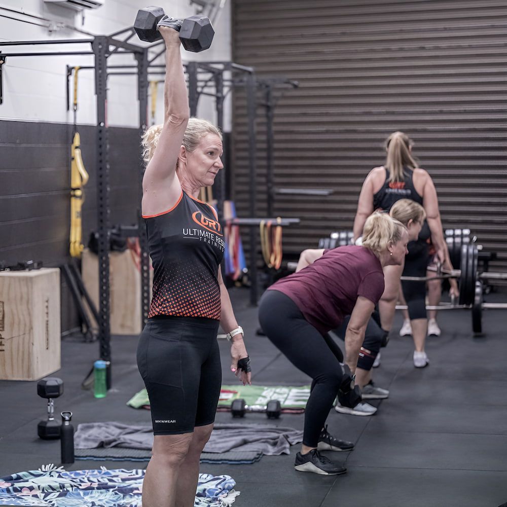 A Woman Is Lifting A Dumbbell Over Her Head In A Gym — Ultimate Results Training in Aitkenvale, QLD